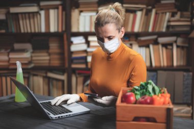Virüs ve patojenlere karşı koruyucu maskeli bir kadın evde dizüstü bilgisayar kullanıyor, internetten yiyecek, yiyecek ve erzak sipariş ediyor..
