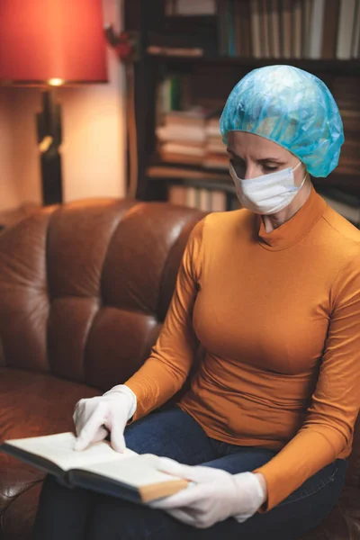 携带防护防病毒面罩的妇女孤零零地坐在家里看书 — 图库照片