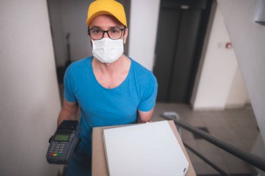 Koruyucu tıbbi maskeli kurye elinde pizza kutusu ve kart ödemeleri için POS kablosuz terminali tutuyor. Virüsler ve salgınlar, evinize yiyecek teslimatı ve güvenlik önlemleri..