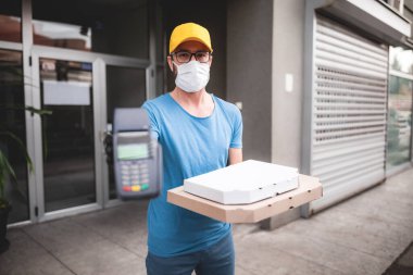 Koruyucu tıbbi maskeli kurye elinde pizza kutusu ve kart ödemeleri için POS kablosuz terminali tutuyor. Virüsler ve salgınlar, evinize yiyecek teslimatı ve güvenlik önlemleri..