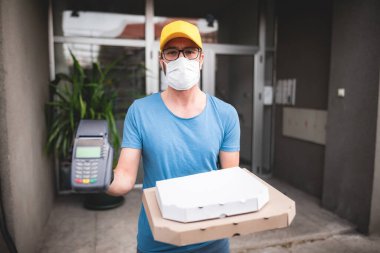Koruyucu tıbbi maskeli kurye elinde pizza kutusu ve kart ödemeleri için POS kablosuz terminali tutuyor. Virüsler ve salgınlar, evinize yiyecek teslimatı ve güvenlik önlemleri..