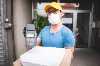 Koruyucu tıbbi maskeli kurye elinde pizza kutusu ve kart ödemeleri için POS kablosuz terminali tutuyor. Virüsler ve salgınlar, evinize yiyecek teslimatı ve güvenlik önlemleri..