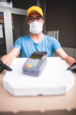 Koruyucu tıbbi maskeli kurye elinde pizza kutusu ve kart ödemeleri için POS kablosuz terminali tutuyor. Virüsler ve salgınlar, evinize yiyecek teslimatı ve güvenlik önlemleri..