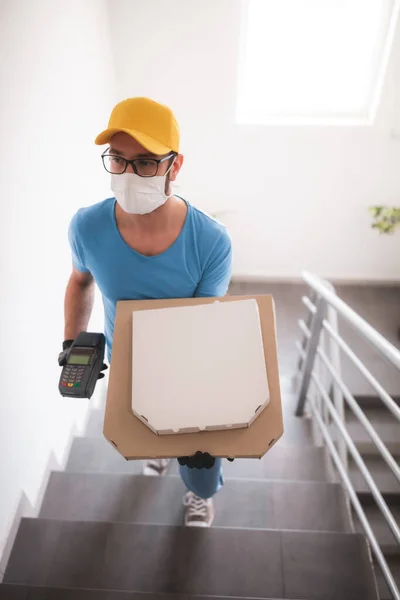 Deliveryman with protective medical mask holding pizza box and POS wireless terminal for card paying - days of viruses and pandemic, food delivery to your home and safety hygiene measures.