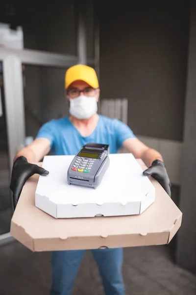 Koruyucu tıbbi maskeli kurye elinde pizza kutusu ve kart ödemeleri için POS kablosuz terminali tutuyor. Virüsler ve salgınlar, evinize yiyecek teslimatı ve güvenlik önlemleri..