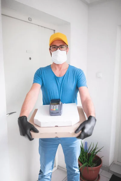 Deliveryman with protective medical mask holding pizza box and POS wireless terminal for card paying - days of viruses and pandemic, food delivery to your home and safety hygiene measures.