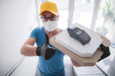 Koruyucu tıbbi maskeli kurye elinde pizza kutusu ve kart ödemesi için POS kablosuz terminali tutuyor. Virüsler ve salgınlarla dolu günler, evinize yiyecek teslimatı..