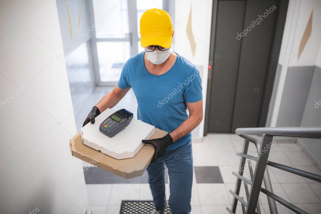 Deliveryman with protective medical mask holding pizza box and POS wireless terminal for card paying - days of viruses and pandemic, food delivery to your home.
