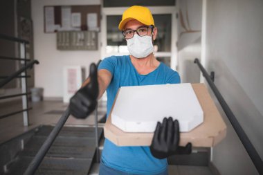 Koruyucu tıbbi maskeli teslimatçı elinde pizza kutusu tutuyordu. Virüsler ve salgınlarla dolu günler, evinize yiyecek teslimatı..