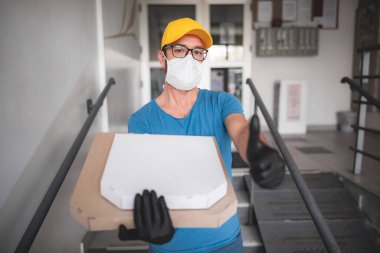 Koruyucu tıbbi maskeli teslimatçı elinde pizza kutusu tutuyordu. Virüsler ve salgınlarla dolu günler, evinize yiyecek teslimatı..