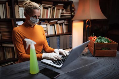Virüs ve patojenlere karşı koruyucu maskeli bir kadın evde dizüstü bilgisayar kullanıyor, internetten yiyecek, yiyecek ve erzak sipariş ediyor..