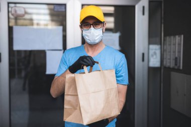 Koruyucu maskeli ve eldivenli teslimatçı. Bir binanın önünde poşet dolusu erzak tutuyor..