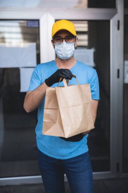 Koruyucu maskeli ve eldivenli teslimatçı. Bir binanın önünde poşet dolusu erzak tutuyor..
