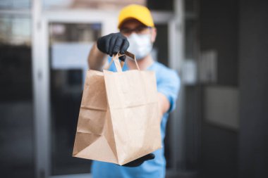 Koruyucu maskeli ve eldivenli teslimatçı. Bir binanın önünde poşet dolusu erzak tutuyor..