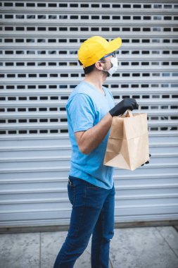 Koruyucu maskeli ve eldivenli teslimatçı. Bir binanın önünde poşet dolusu erzak tutuyor..