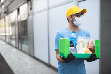 Koruyucu maskeli teslimatçı. Bakkaliye ve POS 'la dolu bir çanta tutuyor..