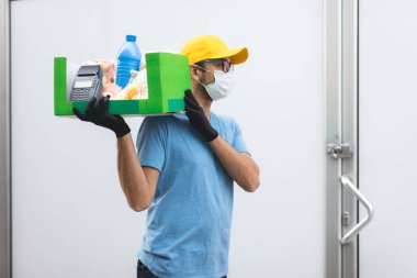 Koruyucu maskeli teslimatçı. Bakkaliye ve POS 'la dolu bir çanta tutuyor..