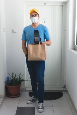 Koruyucu maskeli teslimatçı. Bakkaliye ve POS 'la dolu bir çanta tutuyor..