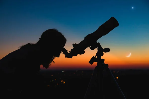 Silhouette of a woman and telescope with twilight sky.