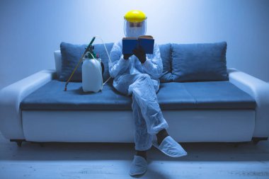 Koruyucu antiviral maske, kostüm, kask ve kimyasal dezenfekte spreyini evde izolasyon şişesinde giyen kişi okuyor..