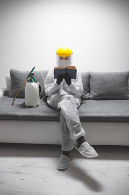Koruyucu antiviral maske, kostüm, kask ve kimyasal dezenfekte spreyini evde izolasyon şişesinde giyen kişi okuyor..