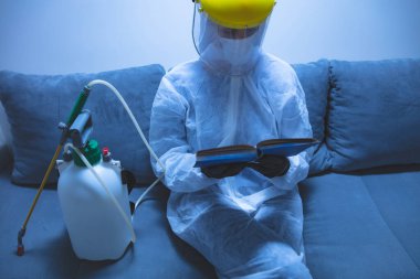 Koruyucu antiviral maske, kostüm, kask ve kimyasal dezenfekte spreyini evde izolasyon şişesinde giyen kişi okuyor..