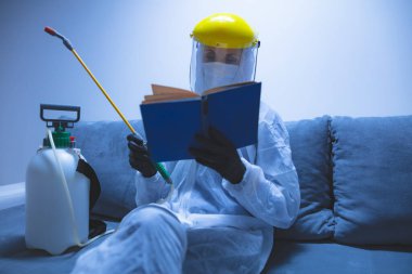 Koruyucu antiviral maske, kostüm, kask ve kimyasal dezenfekte spreyini evde izolasyon şişesinde giyen kişi okuyor..