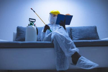 Koruyucu antiviral maske, kostüm, kask ve kimyasal dezenfekte spreyini evde izolasyon şişesinde giyen kişi okuyor..