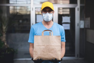 Koruyucu maskeli ve eldivenli teslimatçı. Bir binanın önünde poşet dolusu erzak tutuyor..