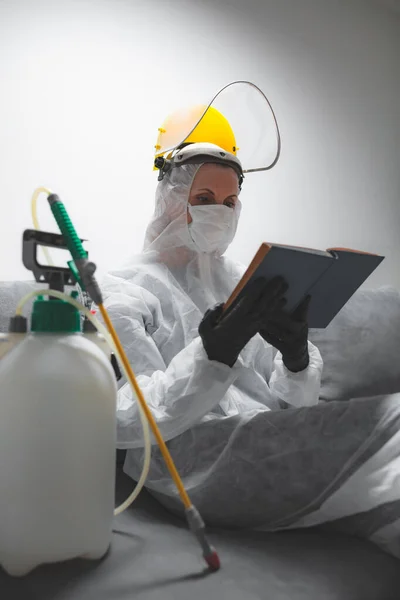 保護抗ウイルスマスク スーツ ヘルメット 家庭用隔離の化学除染スプレーボトルを身に着けている本を読む者 — ストック写真