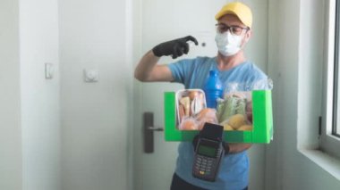 Koruyucu maskeli teslimatçı. Bakkaliye ve POS 'la dolu bir çanta tutuyor..