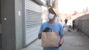 Koruyucu maskeli ve eldivenli teslimatçı. Bir binanın önünde poşet dolusu erzak tutuyor..