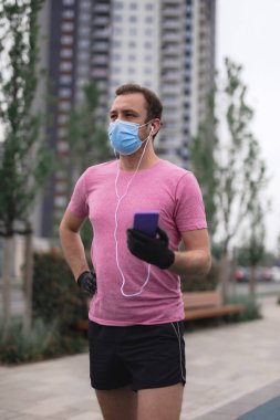 Tıbbi maskeli, eldivenli, akıllı telefonlu ve kulaklıklı sporcu şehir içinde koşuyor.. 