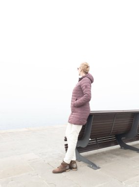 Yalnız bir bankta umutsuzluk içindeki bir kadın ve sisli bir okyanus gününde gri bir ufka bakıyor..