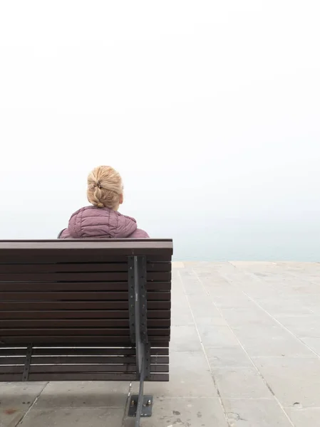 Woman in despair sitting on a lonely bench and looking at grey distant horizon on a ocean foggy day.