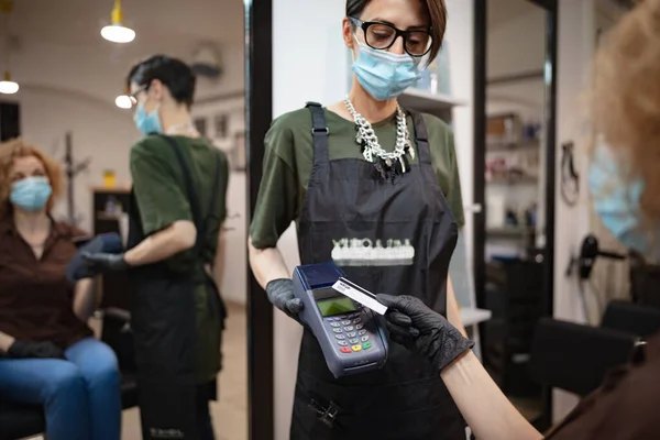 Paying with contactless card. Hairdresser and customer in a salon with medical masks during virus pandemic. Working with safety mask.