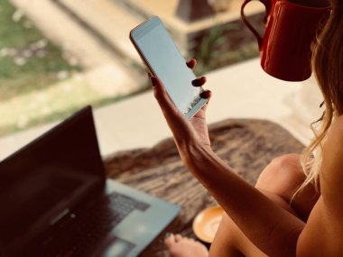 Modern serbest çalışan genç bir kadın cep telefonu / laptop kullanıyor ve teras kanepesinde kahve içiyor..