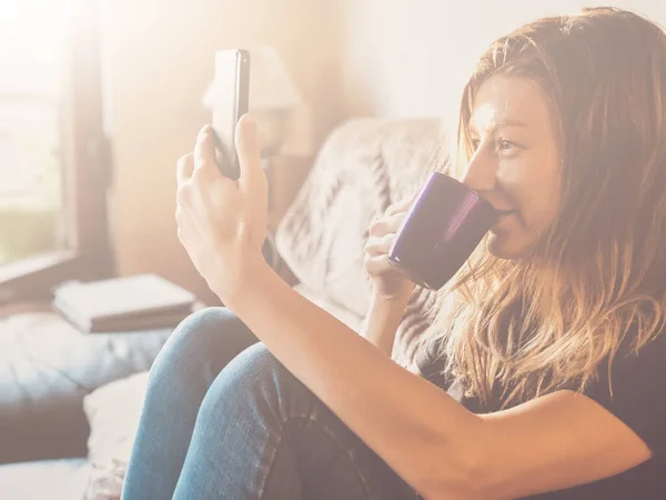 Woman using cellphone and drinking coffee alone at home - ordering food and groceries from home, over the phone during isolation.