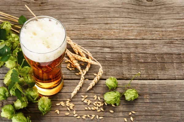 Glass beer glass with foam with hops cones, barley grains and wheat ears on dark wooden table background with copy space, top view