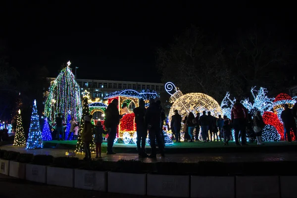 Varna, Bulgaristan - 7 Aralık 2019 - Renkli Noel süslemeleri ve geceleri ışıklar, yeni yıl tatili dekorasyonu, Varna şehir meydanı, insanların silüetleri