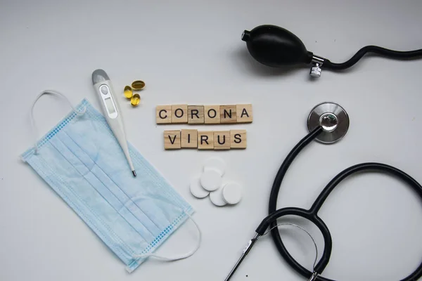 Beyaz arka planda CORONA VIRUS harfli ahşap fayanslar, covid-19, taç virüsü, termometre ve maske, sağlık steteskobu ve kan basıncı ölçer