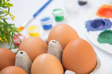 Kutuda paskalya yumurtası, plastik çiçekler ve bulanık boyalar, arka planda renkli paletler, beyaz arka planda boya ekipmanları.