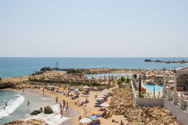 Sahildeki insanlar, sahil şeridindeki lüks otelin yanında eğleniyorlar, havuz, şemsiye ve plaj sandalyeleri.