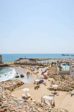 Sahildeki insanlar, sahil şeridindeki lüks otelin yanında eğleniyorlar, havuz, şemsiye ve plaj sandalyeleri.