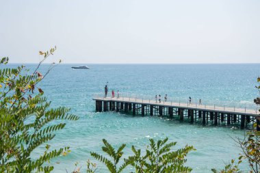 Poseidon heykeliyle iskelede yürüyen insanlar, sahile yakın köprü, mavi deniz ve gökyüzü.