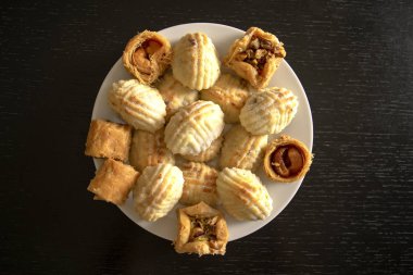 Geleneksel doğu Mamoul kurabiyeleri ve çeşitli baklawa. Üst Manzara. Siyah ahşap masada beyaz tabakta pişmiş Arap tatlısı. Ayd El Fithr ziyafet kurabiyeleri.
