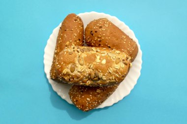 Mavi arka planda beyaz tabakta çok tahıllı mini ekmek. Düz yerde, üst tarafta. Farklı tohumlarla kahverengi ekmek - chia, balkabağı, keten, susam. Küçük somunlar. 