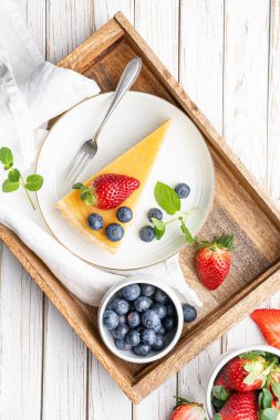 Limon kremalı krem peynirli tart dilimleri, taze yaban mersinleri ve beyaz kırsal arka planda çilek ile servis ediliyor.