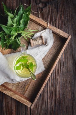 Sağlıklı tıbbi içecek, cam bardakta ısırgan otu çayı, kırsal ahşap arka planda taze yapraklarla süslenmiş.