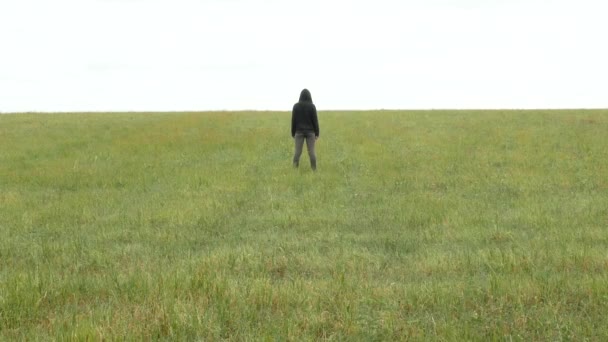 Chica en traje deportivo caqui y capucha se encuentra en un prado en el horizonte — Vídeos de Stock